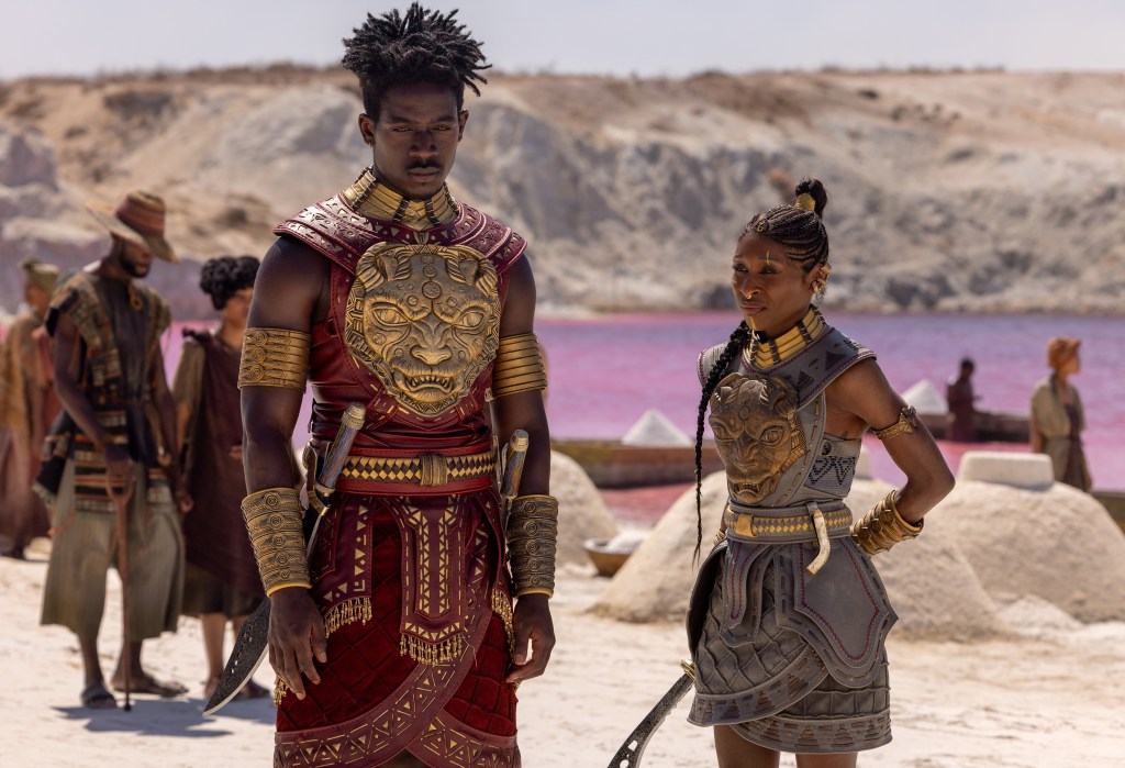 Two people in elaborate tribal costumes stand in a desert landscape with a pink lake in the background.