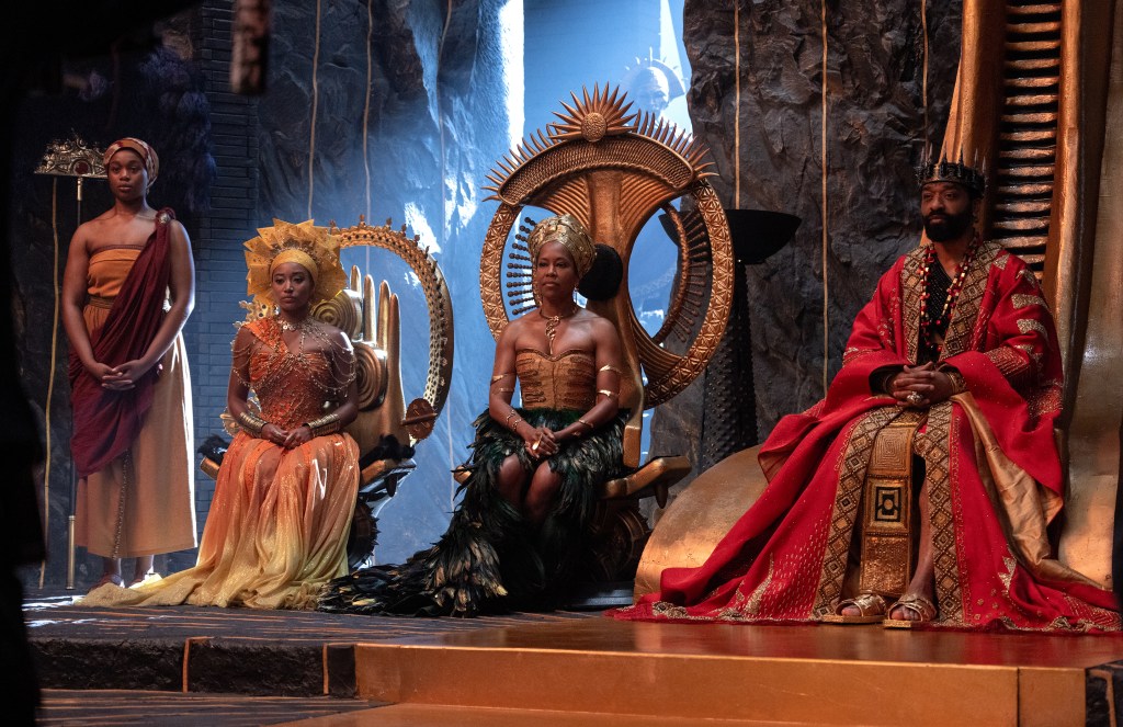 Four individuals in elaborate, colorful costumes and headdresses seated on a throne-like stage with a large ornate mirror behind them.