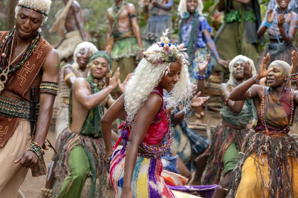 A group of colorfully dressed people, including a woman with long white hair and a flower crown, dancing and celebrating in an outdoor setting.