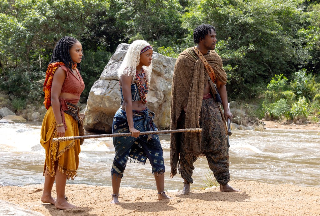 Three people in traditional African attire standing in a river, one holding a staff.