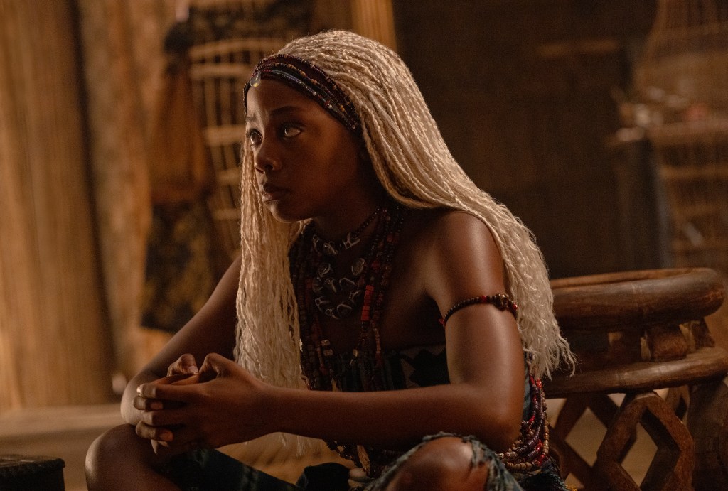 A young woman with long braided hair sits in a dimly lit room, wearing a patterned dress and jewelry. The image has a warm, cinematic tone.