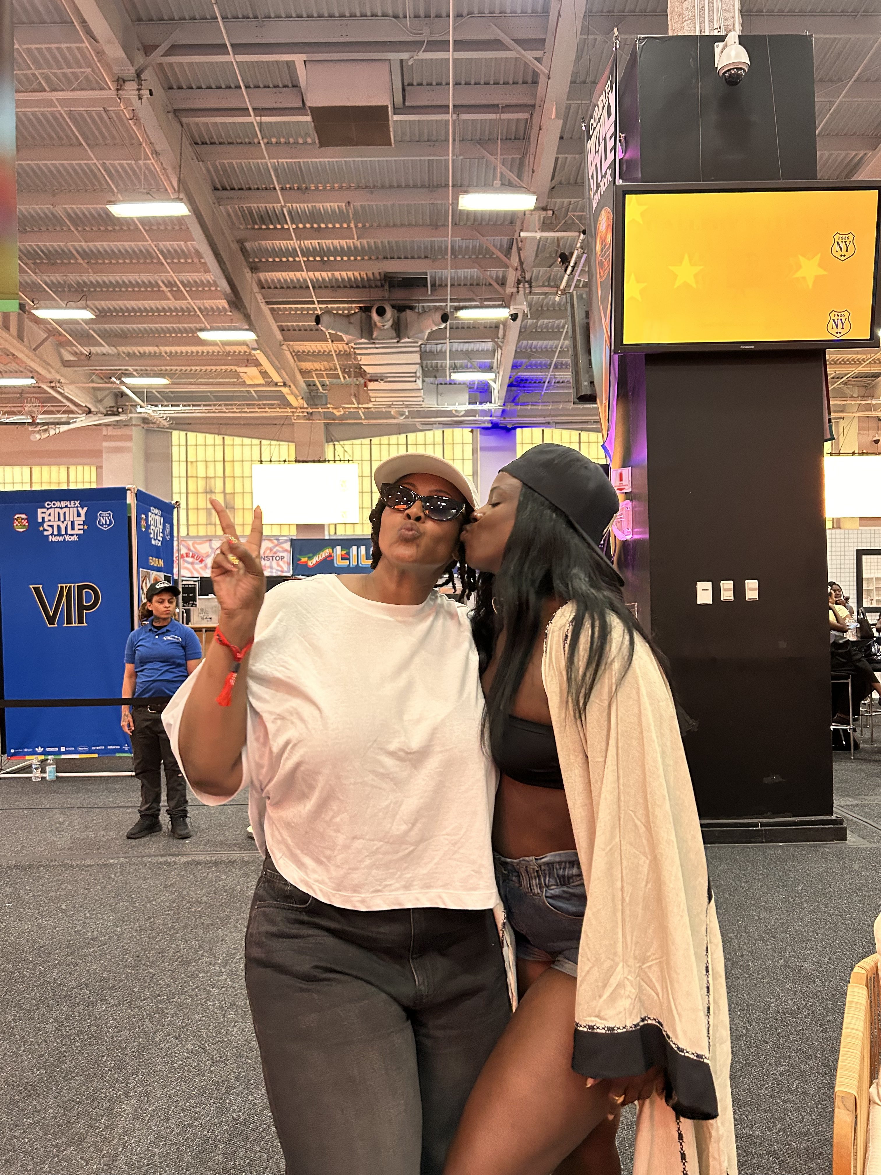 Two people posing for a photo, making peace signs, in front of a large screen displaying a yellow logo.