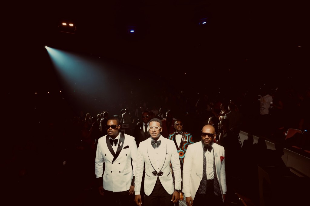 A group of well-dressed men, including three in tuxedos, standing on a stage in front of a large audience.