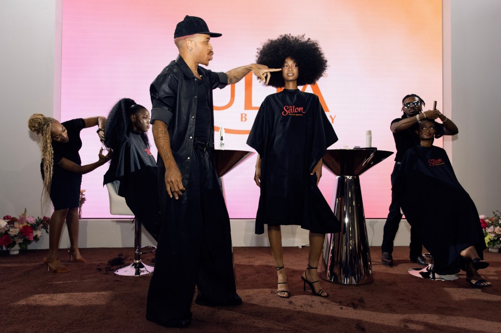 A group of people, including a man in a black hat and a woman with an afro hairstyle, posing in front of a pink and orange backdrop with a "Salon" logo.