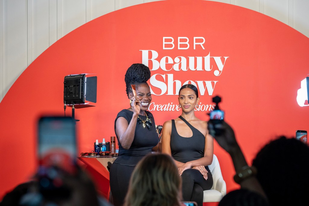 Two women on stage at the BBR Beauty Show, one speaking into a microphone and the other seated next to her.