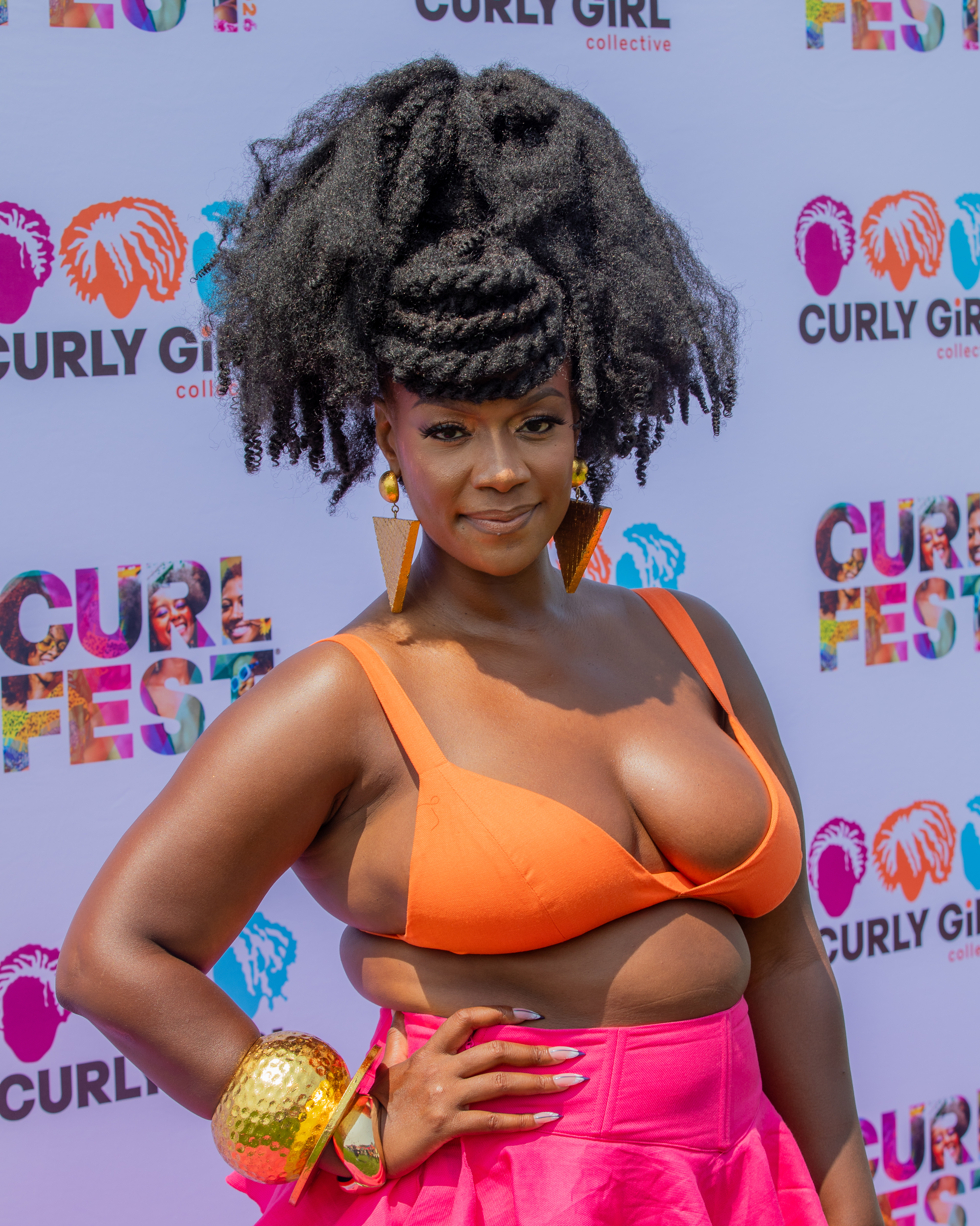 A woman with curly hair wearing an orange top and pink skirt posing in front of a backdrop with the "Curly Girl Collective" logo.